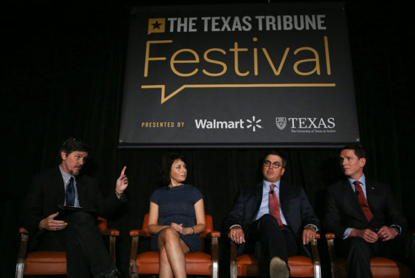 three men wearing business suits and ties and one woman wearing a dress sit in chairs below a large banner that says the texas tribune festival in yellow and white letters with black background