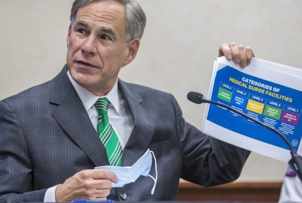 Texas Governor Greg Abbott sitting in front of a microphone holding a chart and face mask.
