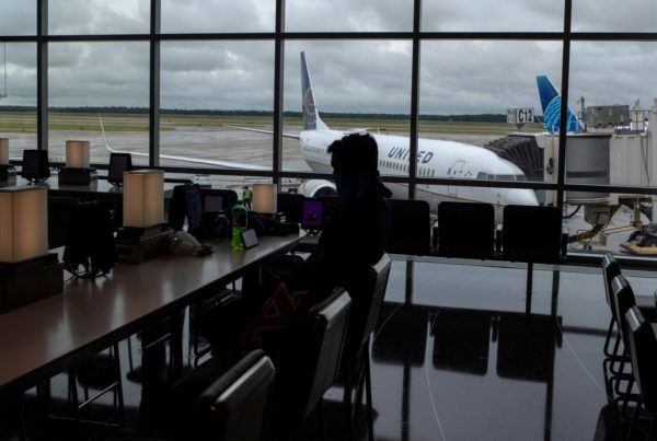 A man sitting in an airport terminal with a plane outside the window behind him.