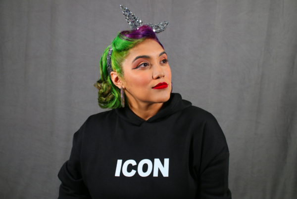 a woman with glamorous makeup, green and purple hair and sparkly siver headband, and black sweatshirt that says Icon