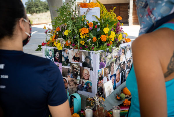 two women stand in front of a box with images of people who've passed away this year with flowers on top