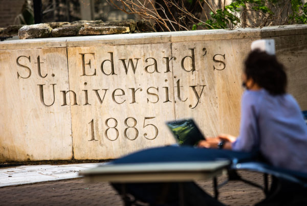 a woman sitting in front of a laptop in front of a sign for st. edward's university