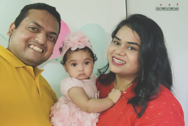 a husband and wife with their baby. he's wearing a yellow shirt, she's wearing a red shirt and the baby is in a pink dress