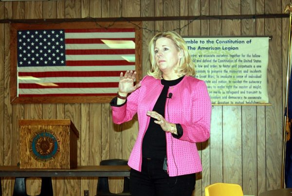 politician Liz Cheney wearing a black outfit and pink jacket speaking at an event in front of an american flag