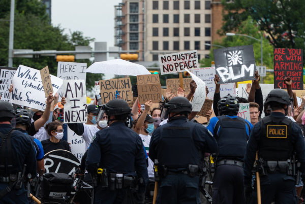 a line of police officers with their backs to the camera, facing protesters with signs