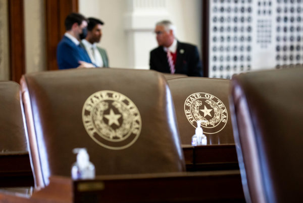 Texas House members converse in the chamber, standing behind empty chairs with the Texas state seal on them.