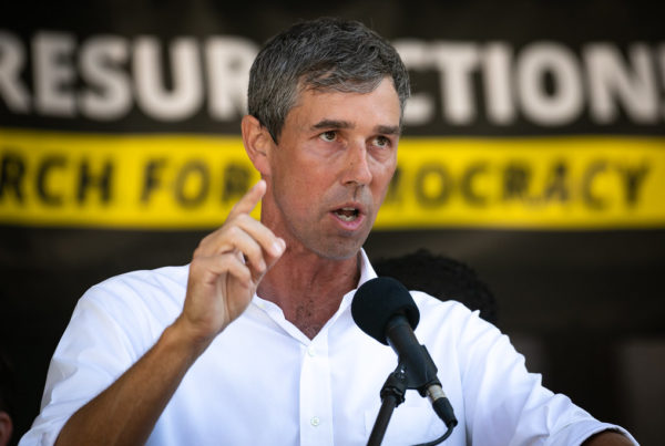 texas gubernatorial candidate beto o'rourke standing in front of a microphone pointing, mid-speec, with a banner in the background that says resurrection and march for democracy