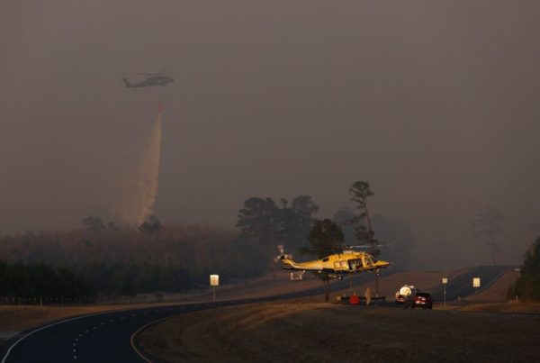a helicopter in the distance dumps water onto a wildfire in bastrop, while another helicopter sits in the foreground