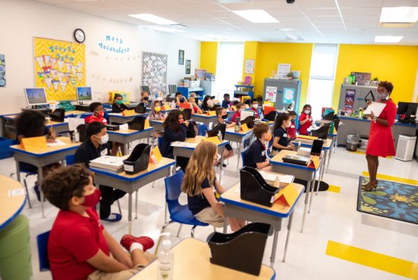 a classroom in houson with bright yellow and white walls, several students looking at a teacher who's wearing a red dress
