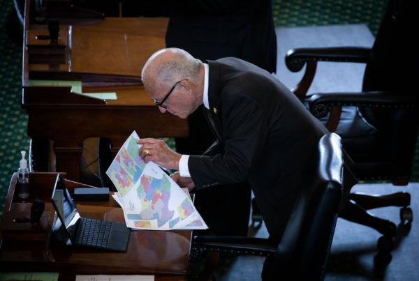 a texas state sematpr wearing a black suit and black glasses bending over a desk looking at a colorful map of political districts, pointing at the map with his finger