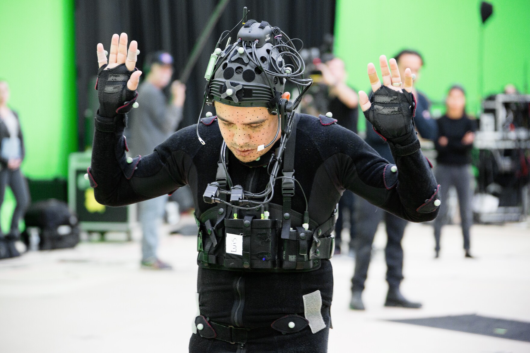 a man wearing black clothes, a helmet and a device over his shoulders and chest to capture his motion, with a green screen in the background