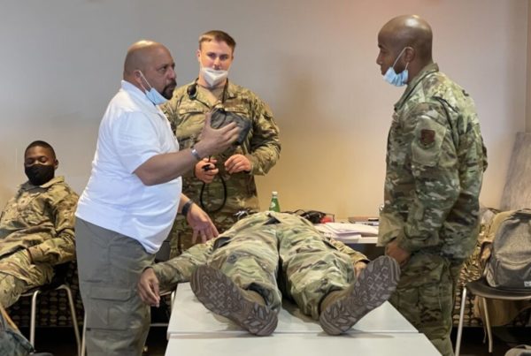 national guard service members in fatigues watch an instructor wearing a white polo shirt instruct them in EMT training while another service member lays on a table in the demonstration