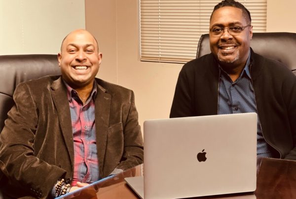 two men wearing button-up shirts and blazers sitting in chairs in front of an apple computer smiling at the camera