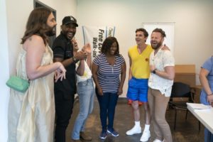 Five people from the cast of the TV show Queer Eye standing around their client Jereka Thomas-Hockaday at her COVID-19 clinic with a banner in the background and some boxes and a chair in the background too