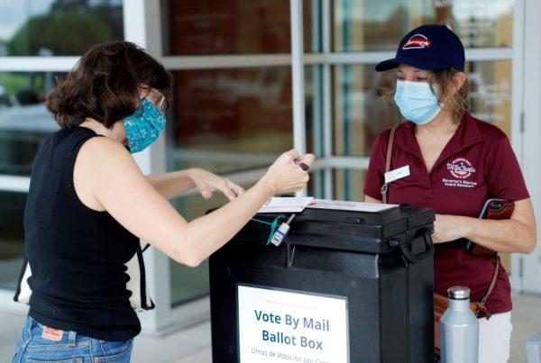 a woman wearing a black top and jeans filling out a form at a vote by mail ballot box while a woman in a red polo t-shirt looks on