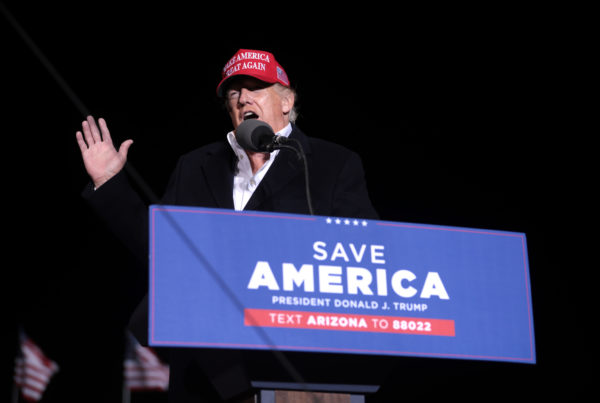 Donald Trump gestures from a podium with the words "Save America" on it. He wears a red hat that says "Make America great again."