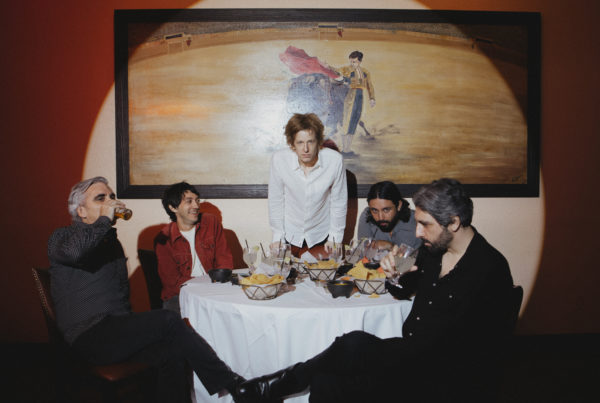 Four band members sit at a table with food, as one member stands at the head.