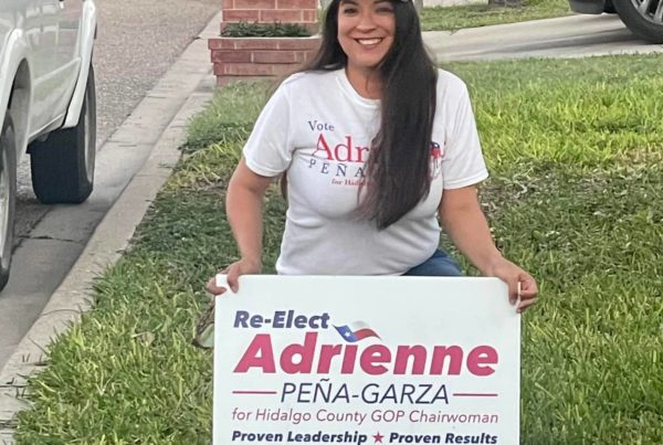 a woman wearing a white hat and t shirt and holding a sign while kneeling on grass that says reelect adrienne pena garza