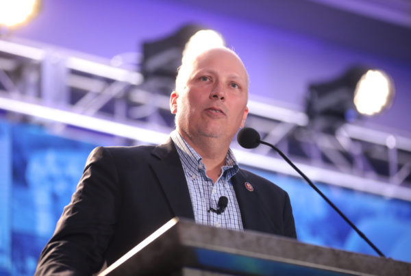 Texas Republican Congressman Chip Roy standing at a lectern with a microphone near his mouth, wearing a dark blazer and checked button-up shirt.