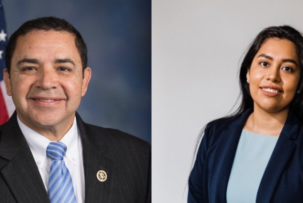 a male politician wearing a blazer and blue tie with an american flag in the background, and a female politician wearing a blue blazer and light blue blouse underneath. she has long dark hair and the background is grey