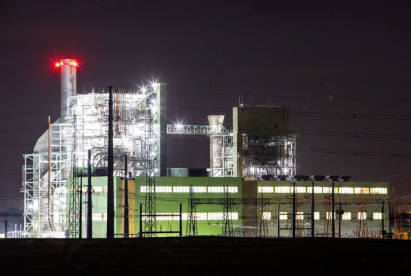a natural gas power plant with glowing white lights and red lights at the top of a large stack, and a long, shorter green building with wide banks of windows on two floors, at night