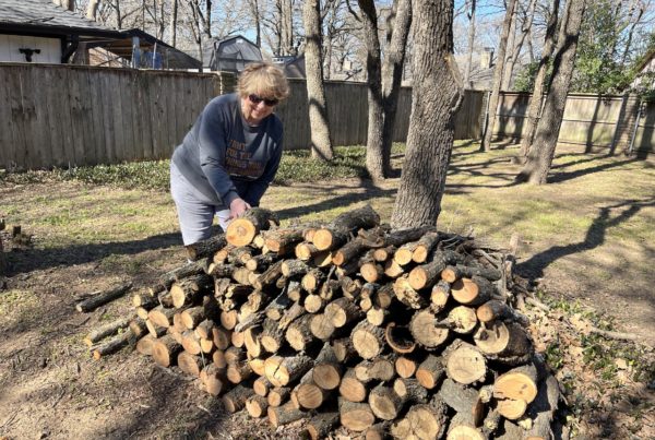 a woman wearing a sweatshirt puts a log on a woodpile outside with trees surrounding her