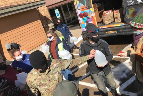 people gather at a water distribution site in Fort Worth as a guy wearing a black sweatshirt, black hat and sunglasses on his head, and a black facemask hands out gallons of water