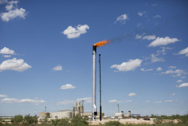 a tall silver pipe with flames coming out of the top with a blue sky and small white clouds in the background