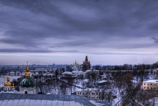 a cityscape of Kyiv, Ukraine with domed buildings, an icy river, and tall tower buildings in the background with a cloudy sky and snow everywhere