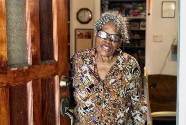 a woman with gray curly hair wearing glasses and a brown purple and other colored blouse standing behind her open front door