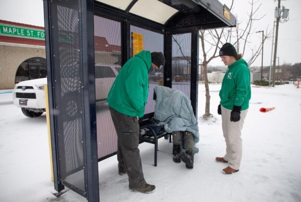 Two people wearing green jackets hats and gloves stand in front of a person experiencing homelessness who's wrapped in a blanket at a bus shelter during a freeze