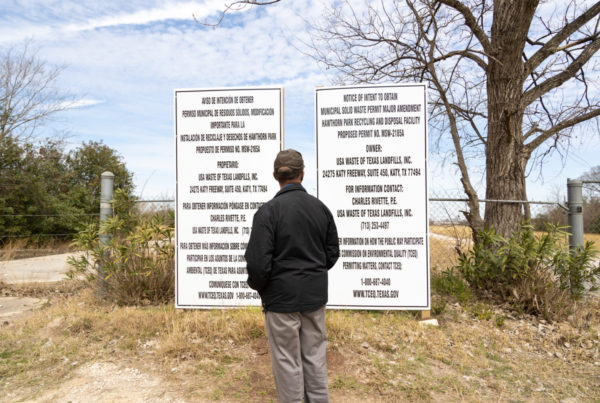 a man wearing a black jacket and baseball cap staring at signs in front of a landfill with trees and bushes in the background