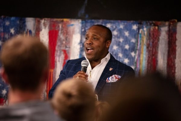 a Black political candidate wearing a blue blazer and white button down shirt holding a microphone and american flag art in the background