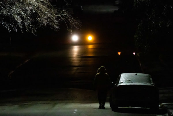 a night photo of a person walking in a winter coat with a car's headlights in the distance and the freezing branches of a tree in the corner