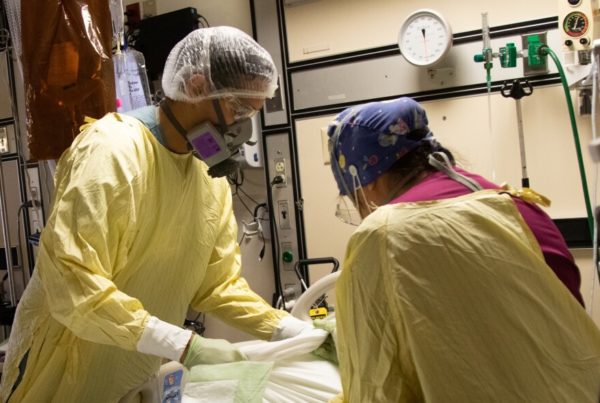 two doctors wearing yellow gowns, caps and face respirators change bed linens on a patient's bed in a hospital