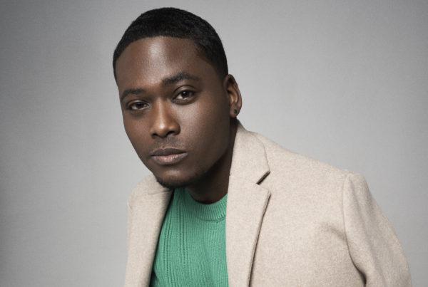 a young man wearing a tan blazer and green sweater looks thoughtfully at the camera