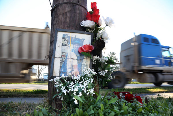 a laminated photo of a young man in a blue graduation cap and gown is secured to a telephone poll next to some red and white flowers as a semi truck passes on the road behind the pole