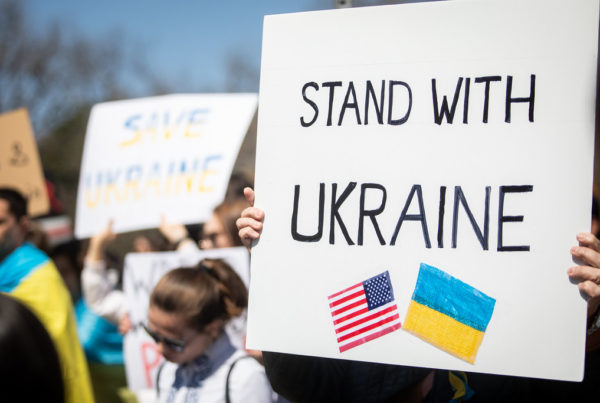 a protestor holds a sign reading "stand with Ukraine" with an American and a Ukrainian flag.