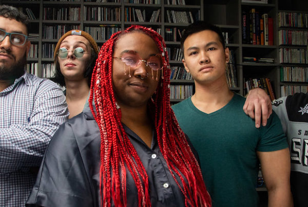 a band with five members, the lead member with bright red braids and glasses with beads haning off the lenses