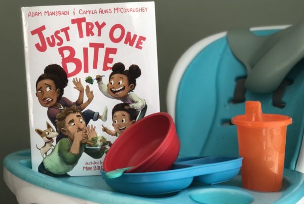 the book "Just Try One Bite" has an illustrated cover with two parents making disgusted faces at three kids trying to offer them vegetables. The book sits on a highchair tray table with a sippy cup and child's bowl and plate.