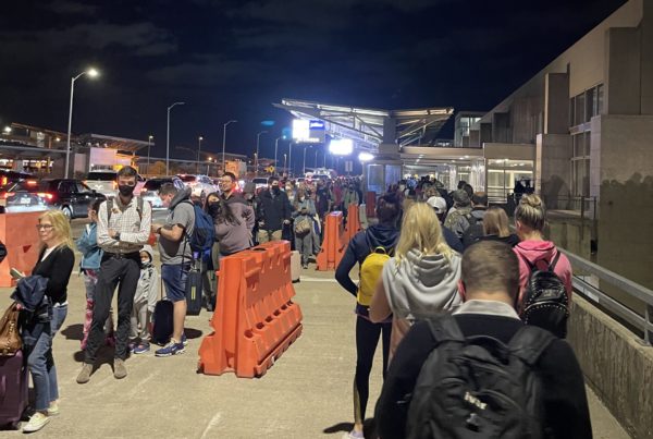 lines of people snake around the sidewalk outside the airport terminal on a dark Monday morning. some are masked, some toting luggage, most checking cell phones.