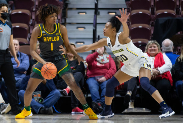 one woman wearing a green and yellow basketball jersey dribbling a ball against another woman player wearing a white jersey holding her arms out