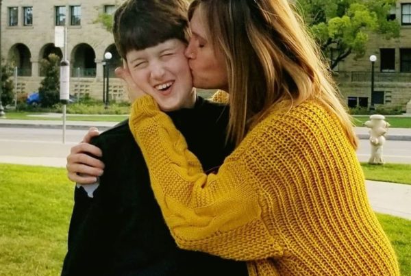 A mom wearing a yellow sweater kisses her child, who's wearing a black jacket, on the cheek