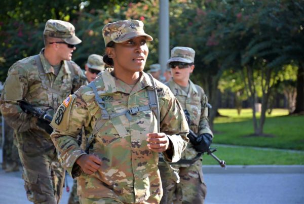army service member carlandra moss wearing fatigues and a camo hat, with other soldiers in the background and trees