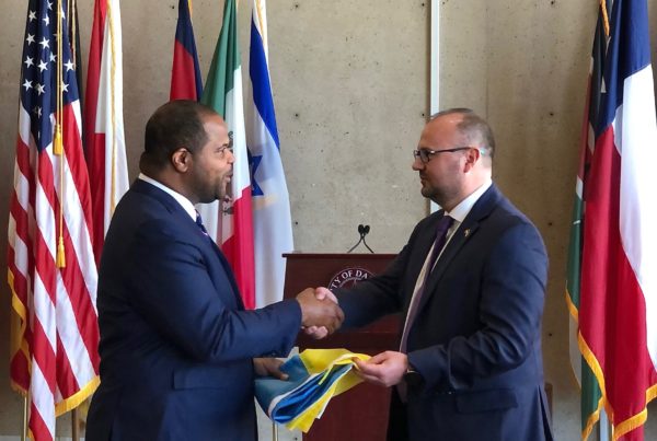two men in blue suits stand facing each other shaking hands in front of several different flags