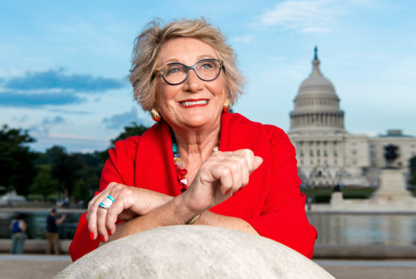 a woman wearing a bright red blazer and glasses perched on a granite carving in the National Mall in front of the U.S. Capitol