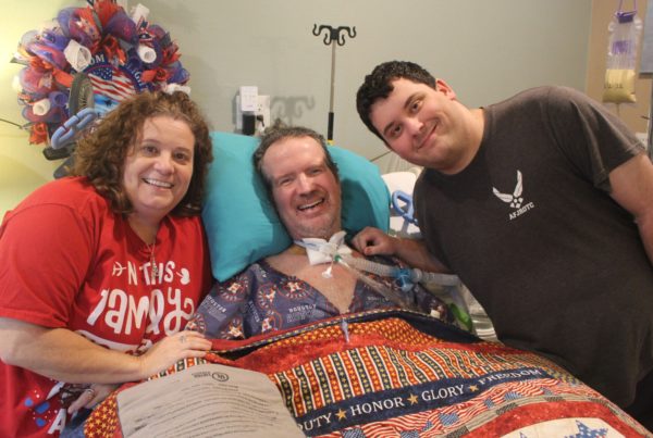 a woman wearing a red t-shirt poses with her husband in the middle, in a hospital bed and a tracheotomy tube, and their son to his right, wearing a black t-shirt