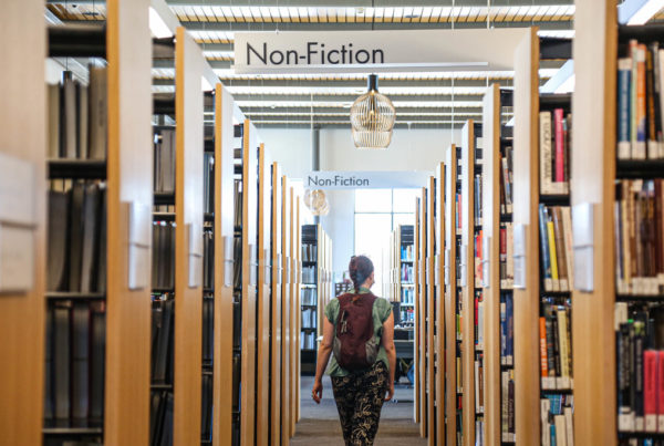 a woman wearing a purple backpack walks down a library aisle with a sign that says nonfiction above her