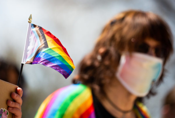 a rainbow flag also with the triangle of blue, pink, white for transgender pride, and black and brown for LGBTQ people of color, waving behind a person wearing a light rainbow face mask with shoulder length brown hair and sunglasses