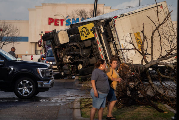 an overturned truck lays on its side with a fallen tree branch in front of a pet smart building with two women looking toward the camera